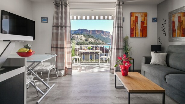 A modern living room with a flat-screen TV, a dining area with a table and chairs, a sofa, and a view of a mountainous landscape. - Apartment 'Le Panorama' with Sea View, Balcony, Wi-Fi and Parking (Cassis)