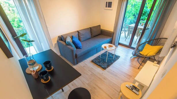 Ein modernes Wohnzimmer mit einem grauen Sofa, einem kleinen runden Tisch und einem schwarzen Sessel. Eine Glastür führt zu einem Außenbereich mit Tisch und Stühlen.