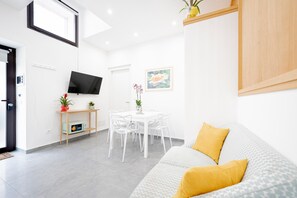 A modern living room with a sofa, a dining table with chairs, a TV, and a small kitchen area. - Two Bedroom Deluxe Apartment (Siracusa)