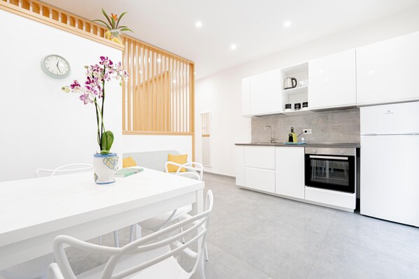 A modern kitchen with white cabinetry, a built-in oven, and a dining area with a white table and chairs. - Two Bedroom Deluxe Apartment (Siracusa)