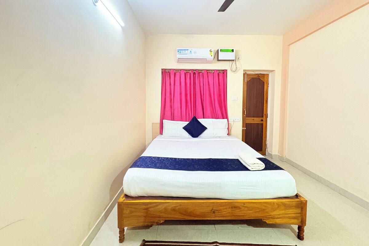 A single bed with a wooden frame, white bedding, and a blue accent pillow. There is a ceiling fan, a wall-mounted air conditioner, and a door with a wooden frame.