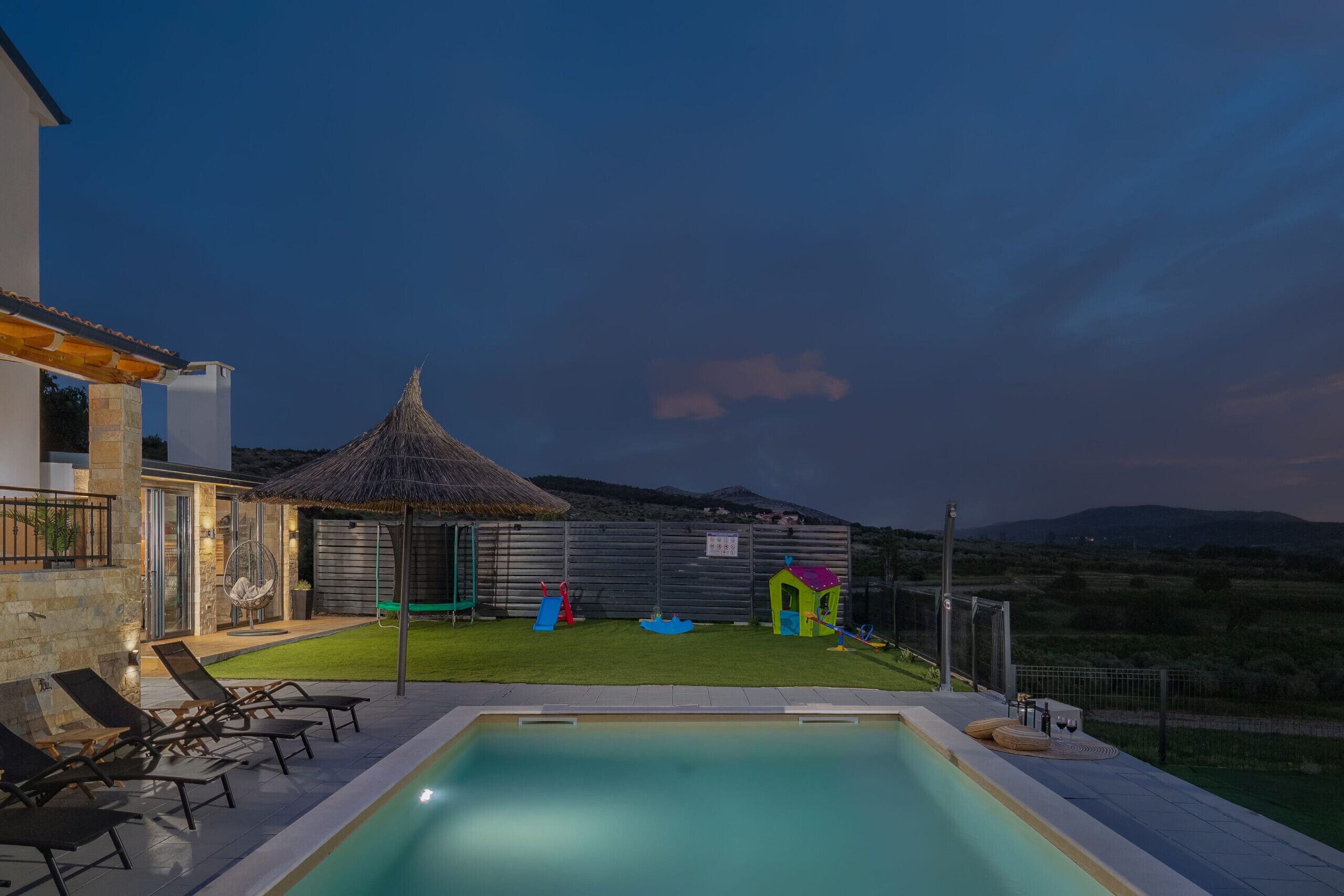 A swimming pool with lounge chairs, a thatched-roof structure, and a fenced play area.