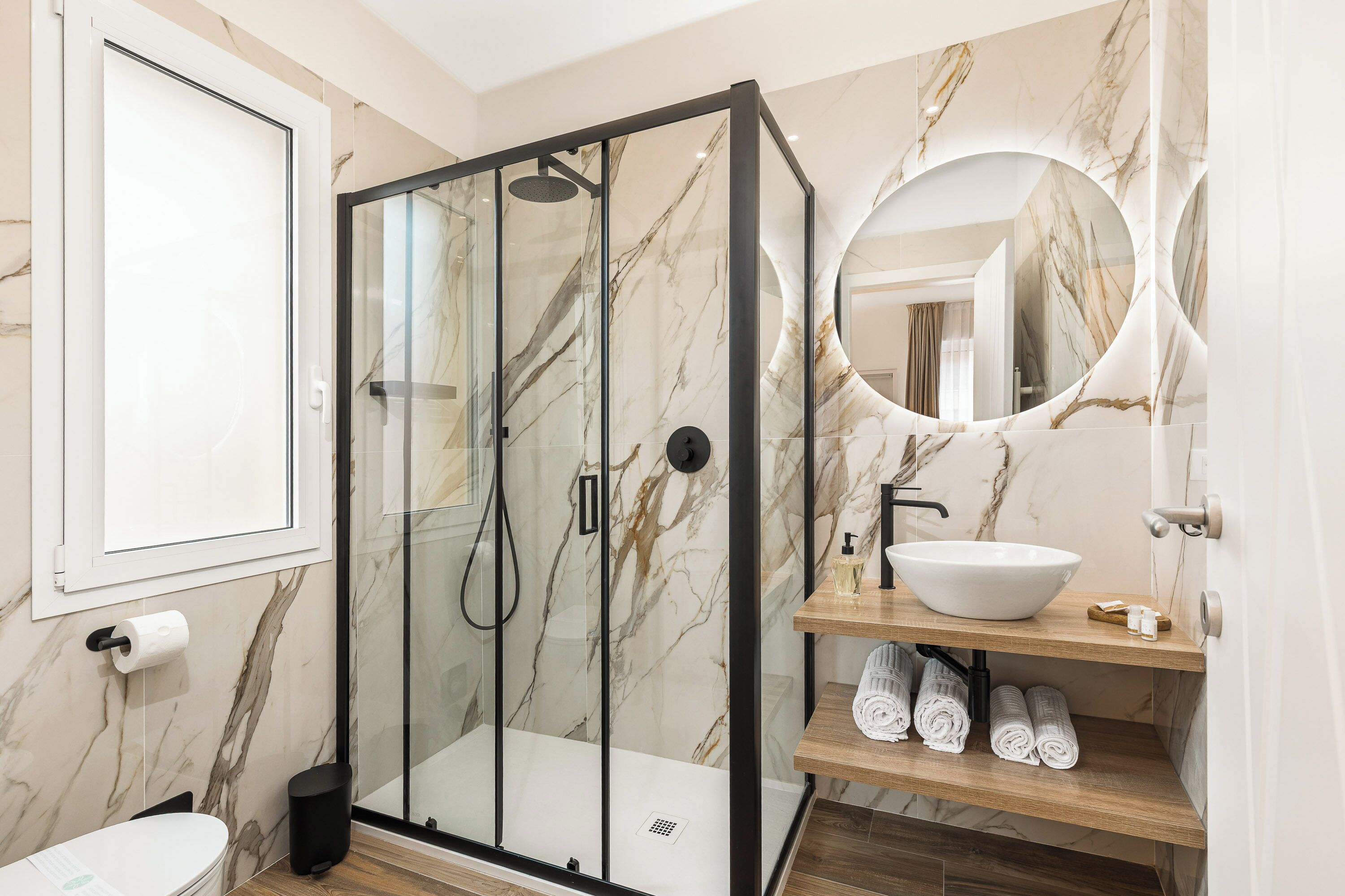 Une salle de bain moderne avec une douche à parois de verre, un mur recouvert de carreaux de marbre, un lavabo blanc et une étagère en bois où sont posées des serviettes.
