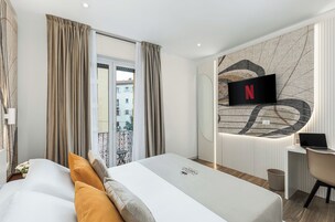 Una camera d'albergo moderna con un letto, una scrivania con un portatile, una TV a parete e un balcone con vista sugli edifici.