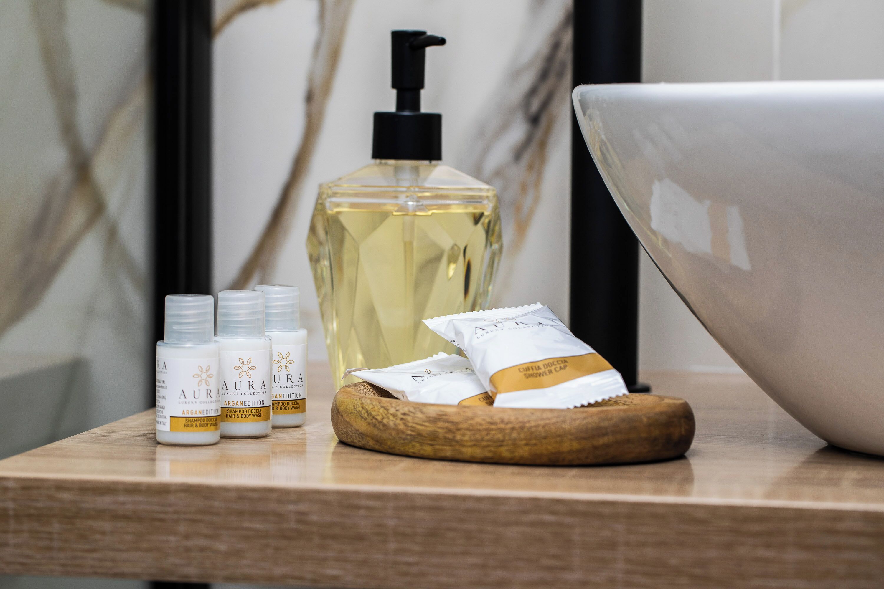 Ein Badezimmer-Waschtisch mit einer Holzschale, einem großen weißen Becken und verschiedenen Pflegeprodukten, darunter eine Lotionflasche und eine Duschhaube.