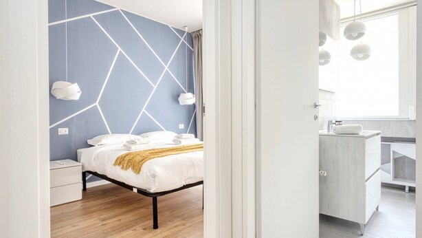 A modern bedroom with a geometric-patterned wall, a bed with white linens, a bedside table, and a nightstand. - Hostmate - Milano - Imperia's Prism - Romolo IULM (Milan)