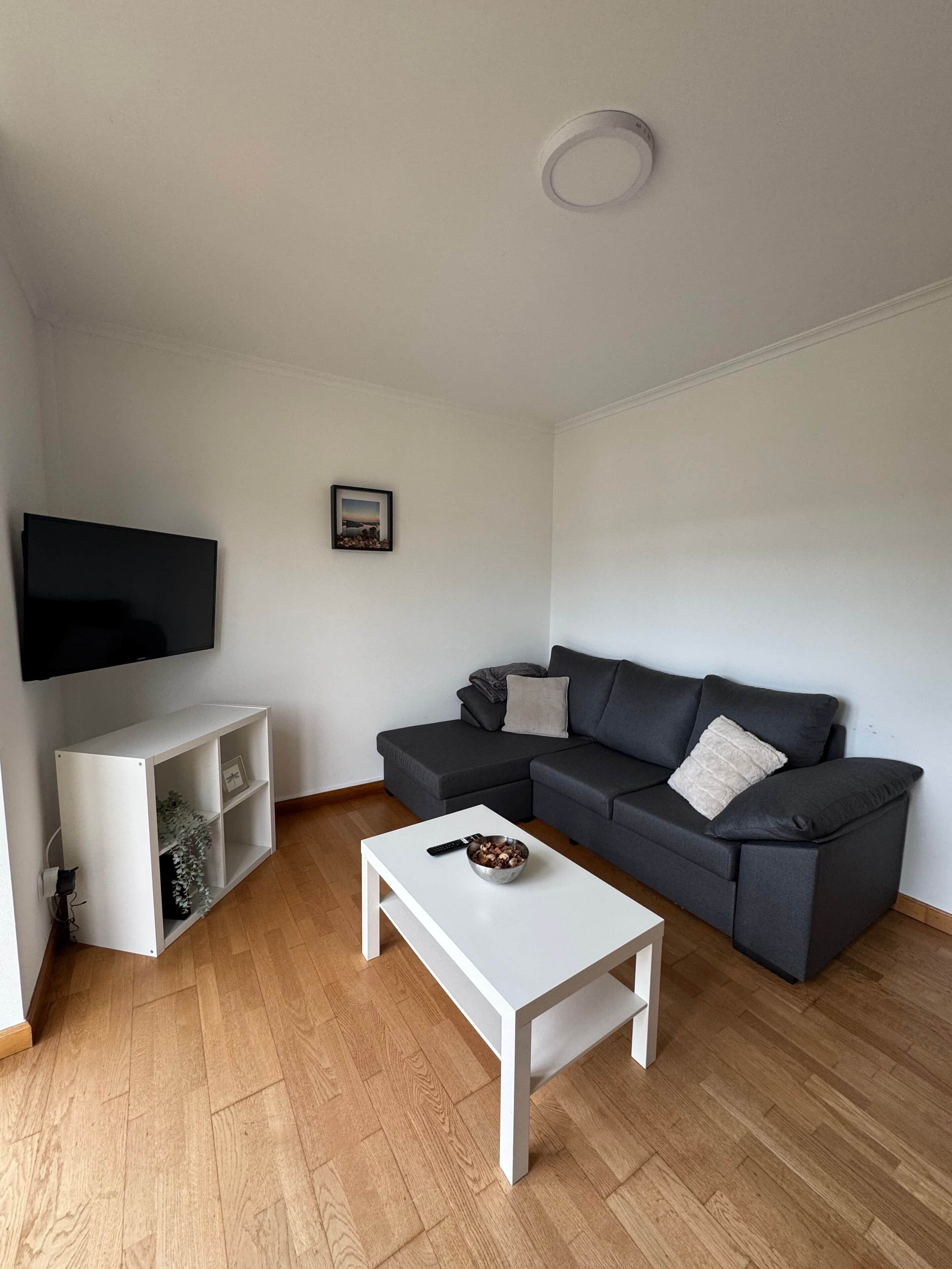 Una sala de estar moderna con un sofá seccional gris, una mesa de centro blanca, un estante blanco y un televisor de pantalla plana montado en la pared.