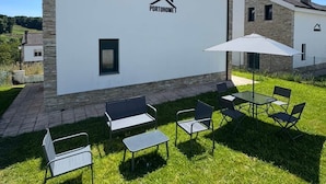 Et moderne hus med hvide facader og stenelementer, med en terrasse med udendørs møbler og stor parasol.