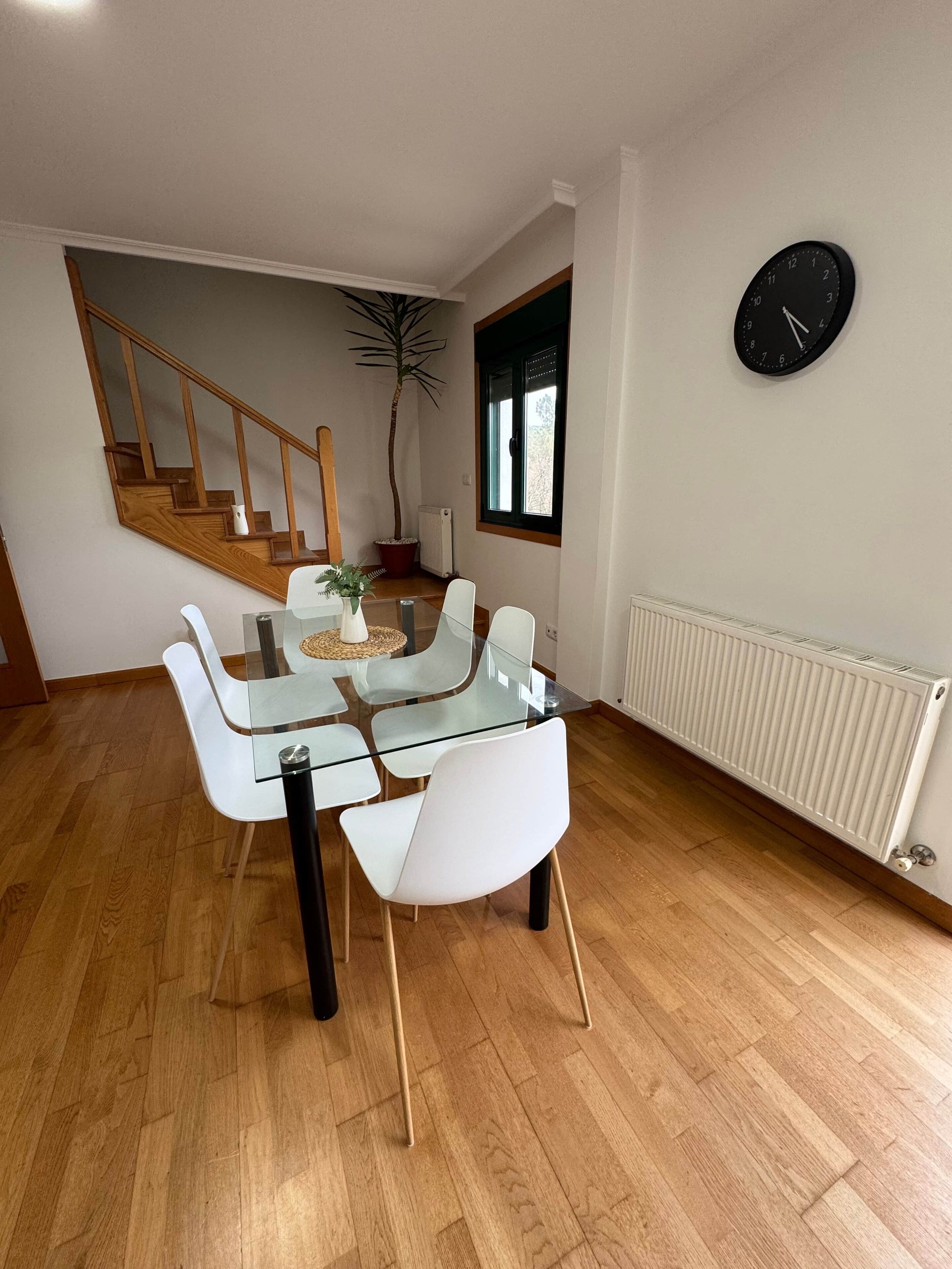 Un comedor con mesa de vidrio y sillas blancas, una escalera, una planta en maceta y un reloj de pared.