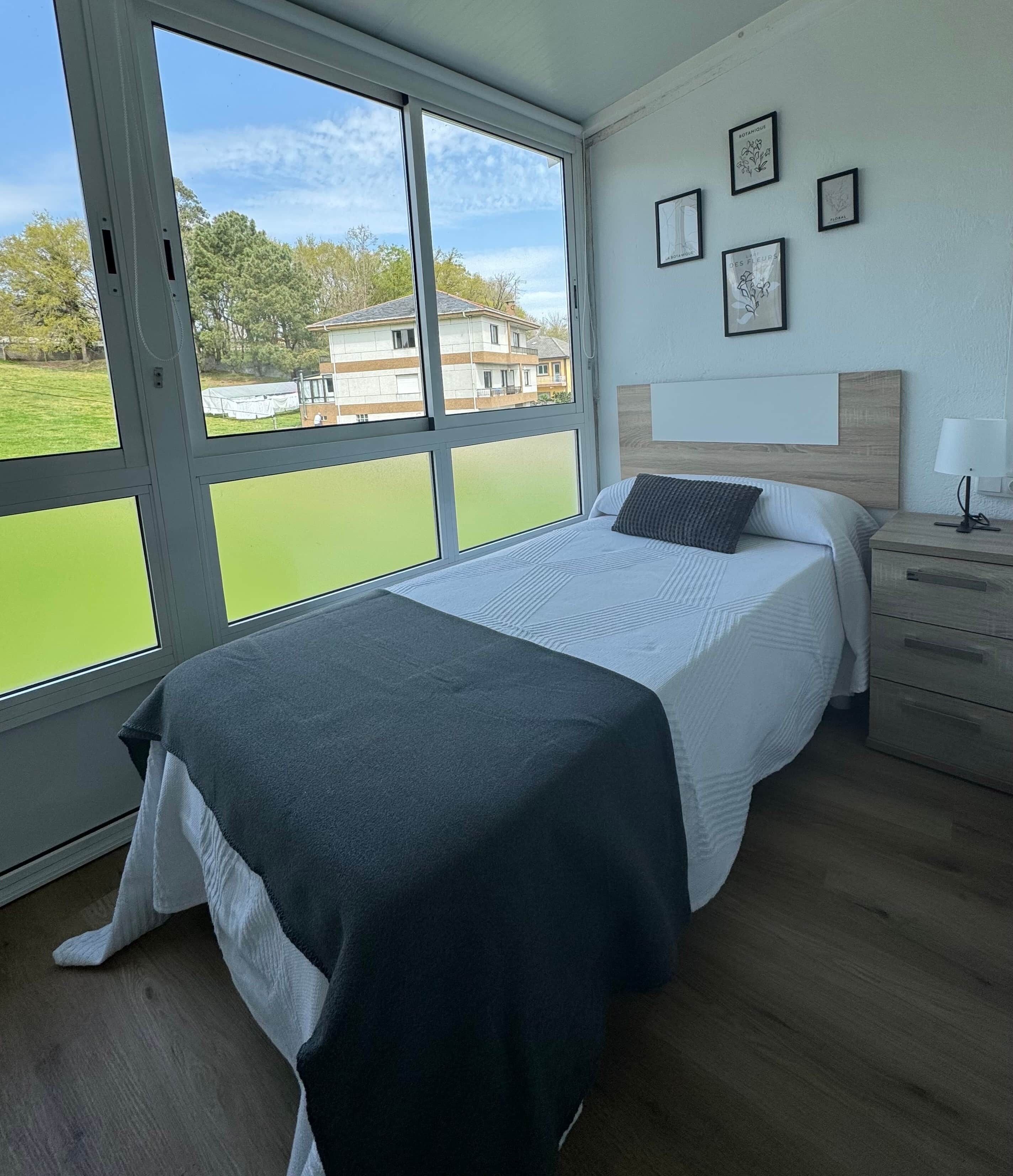 Un dormitorio con cama, mesita de noche, lámpara y vistas a zonas verdes y edificios a través de amplias ventanas.