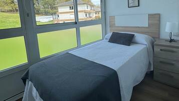 Un dormitorio con cama, mesita de noche, lámpara y vistas a zonas verdes y edificios a través de amplias ventanas.