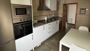 Een moderne keuken met witte kasten, een ingebouwde magnetron, oven en een roestvrijstalen afzuigkap. Er is een eettafel met stoelen en een klein eiland met een spoelbak.
