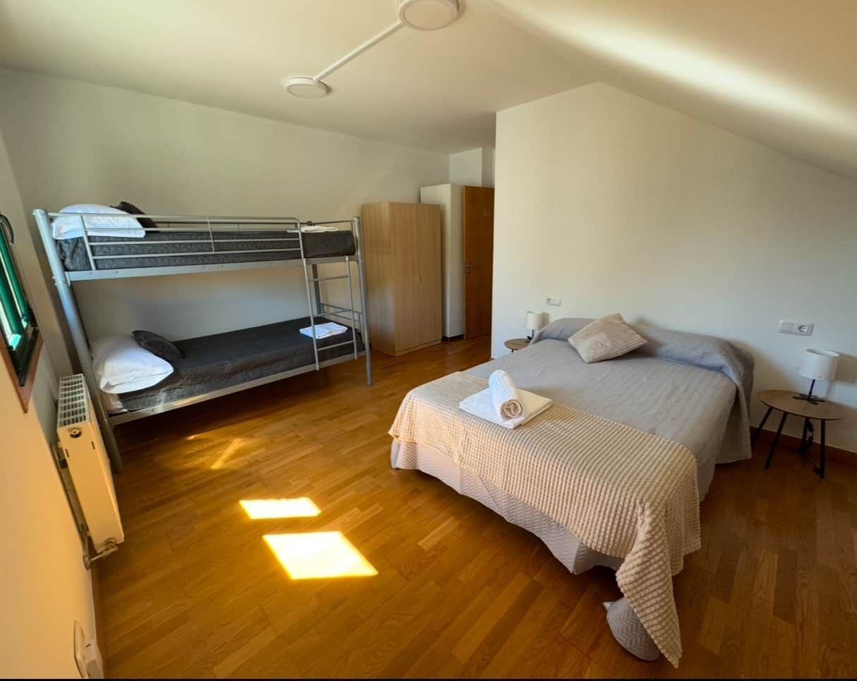 Une chambre avec un lit superposé, un lit simple, un plancher en bois, une armoire et une petite table avec une lampe.