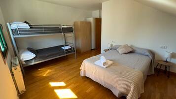 Une chambre avec un lit superposé, un lit simple, un plancher en bois, une armoire et une petite table avec une lampe.