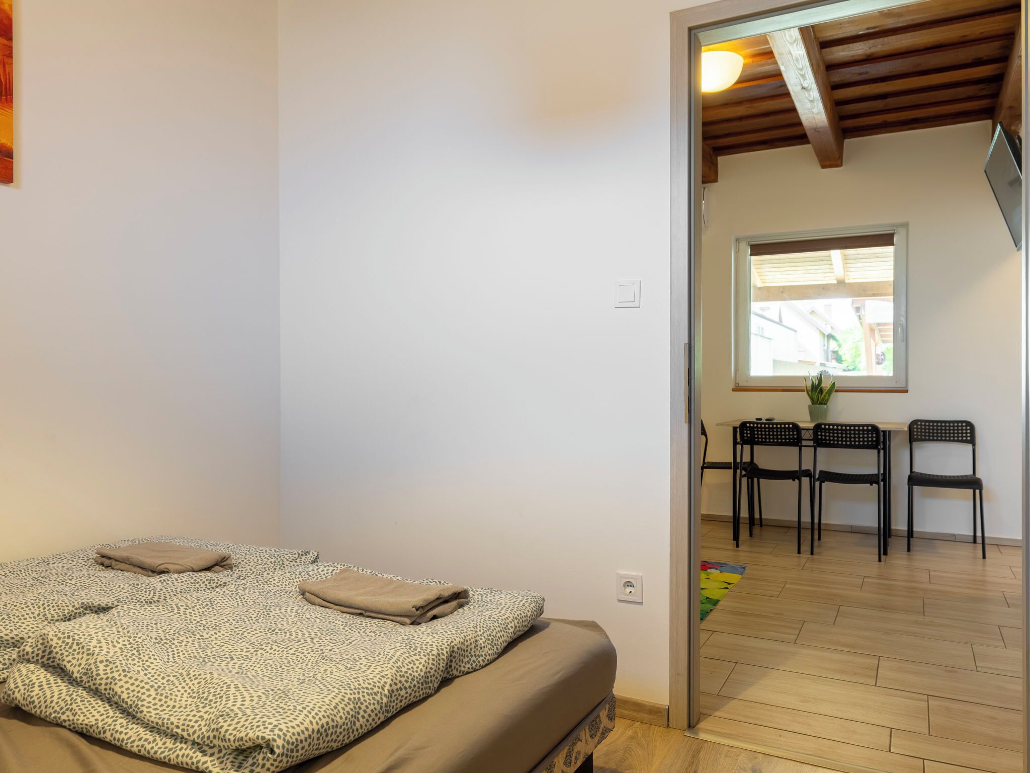 A bedroom with a bed, a small table with chairs, a window, and a wooden ceiling.