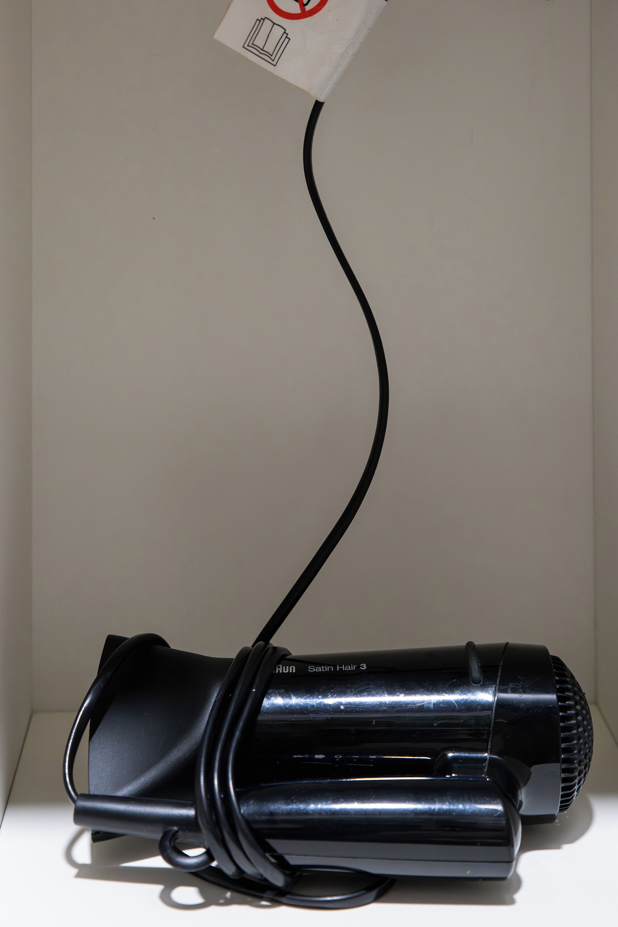 A black hairdryer with a cord plugged in, placed on a white surface against a plain wall.