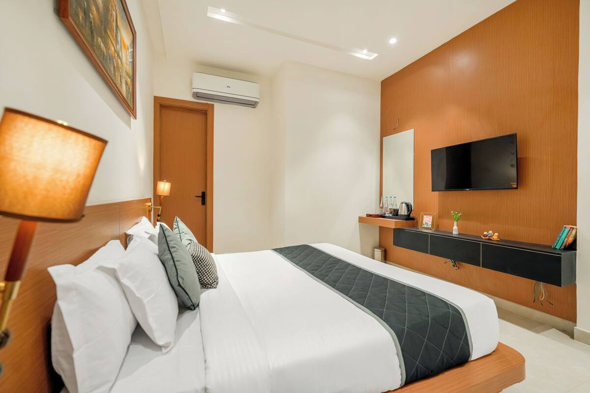A modern hotel room with a neatly made bed, a mounted TV, a wooden wardrobe, and a wall-mounted air conditioner.