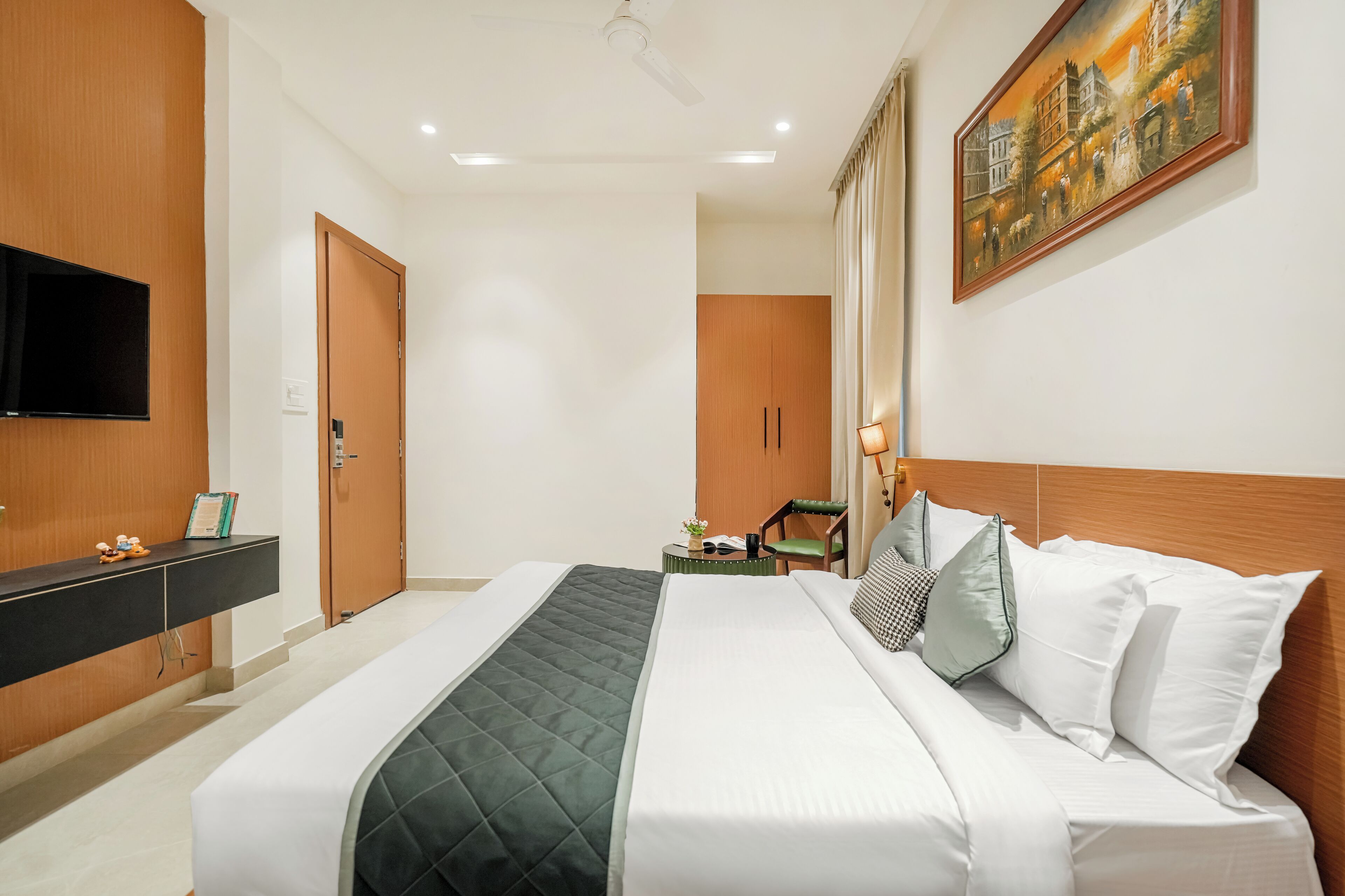 A hotel room with a large bed, a flat-screen TV, a wooden wardrobe, a desk with a chair, and a painting on the wall.