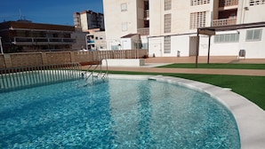 Una piscina cristalina con una escalera de metal, rodeada por un edificio blanco con balcones y un césped verde.