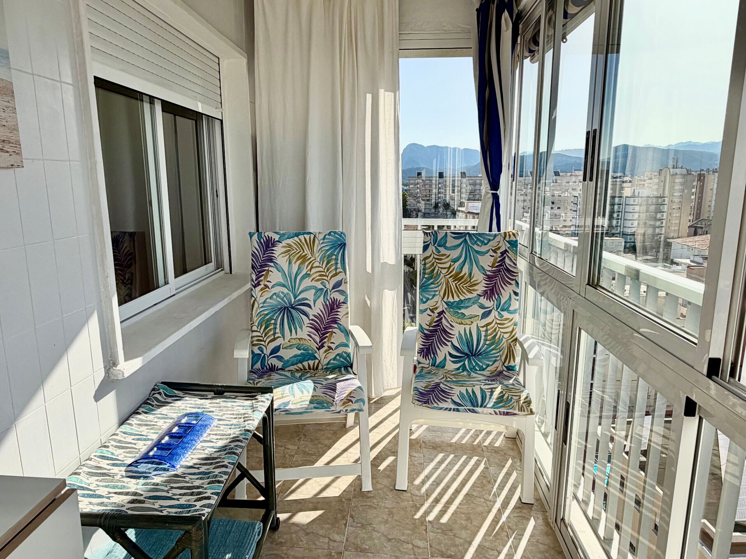 Un balcon avec une table et deux chaises, une vue sur les bâtiments et les montagnes, et une balustrade blanche.