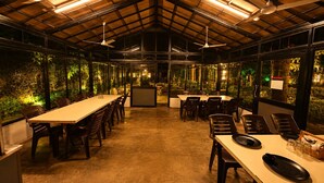 Un salón de conferencias con mesas largas y sillas, ventiladores de techo y ventanas grandes con vistas a la vegetación.