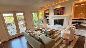 Ein geräumiges Wohnzimmer mit Sofa, Hocker und Sesseln, einem Kamin und einem Bücherregal.