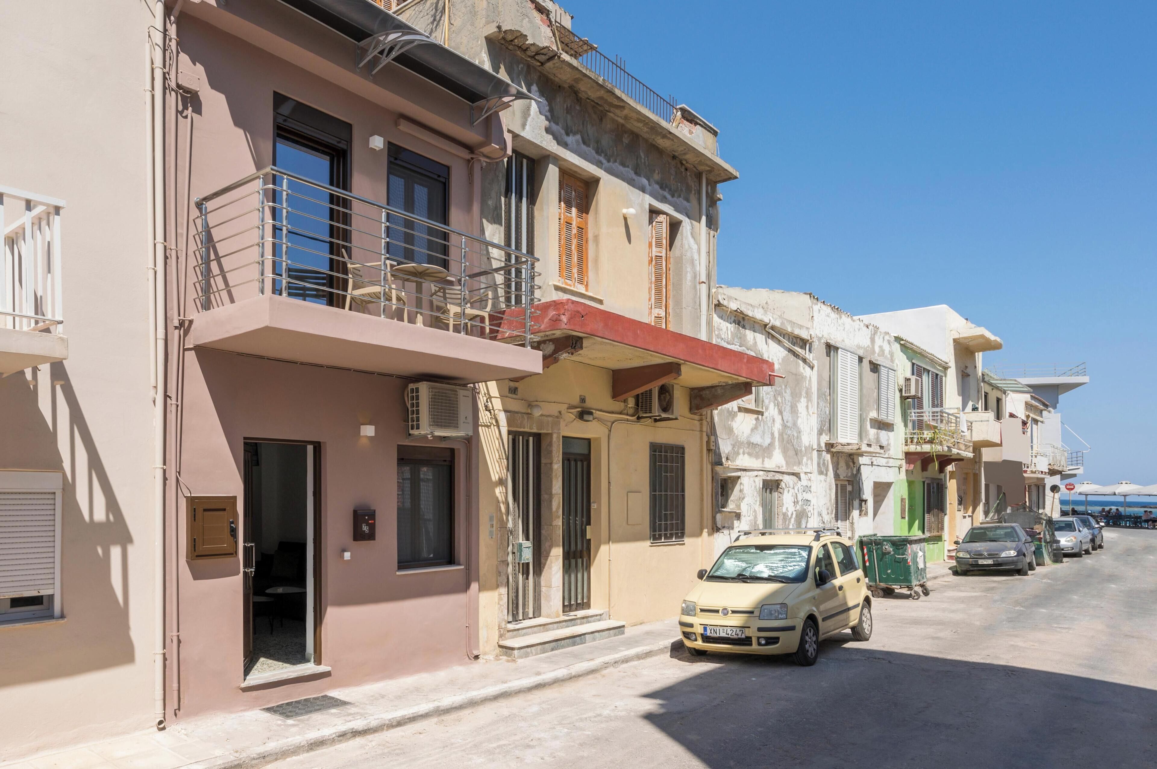 A row of multi-story buildings with balconies, a car parked in front, and a clear blue sky.