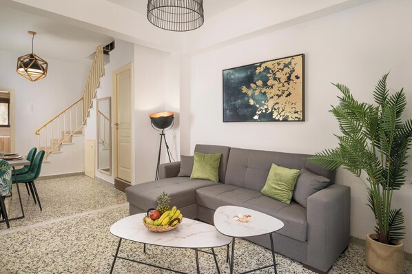 Ein Wohnzimmer mit grauem Sofa, einem weißen Couchtisch, einem Essbereich mit Tisch und Stühlen, einer Treppe und einer großen Pflanze.