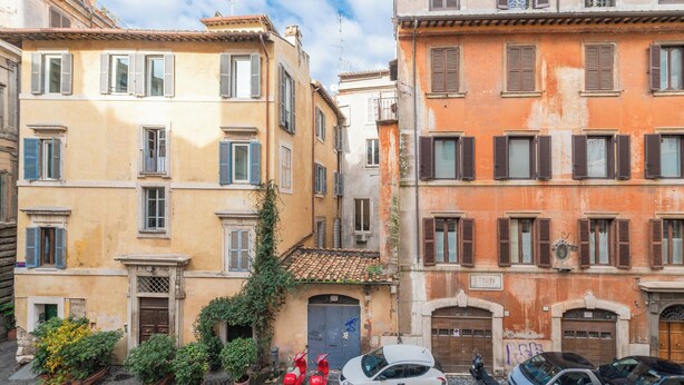 A narrow street with buildings on either side, featuring a row of parked cars and a few potted plants. - Nosdom Properties - Piazza Navona Design Apartment (Roma)