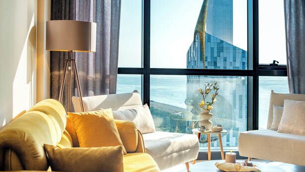 Ein modernes Wohnzimmer mit einer Couch, Sesseln, einem Couchtisch und einer Stehlampe. Ein großes Fenster bietet Blick auf ein hohes Gebäude und das Meer.
