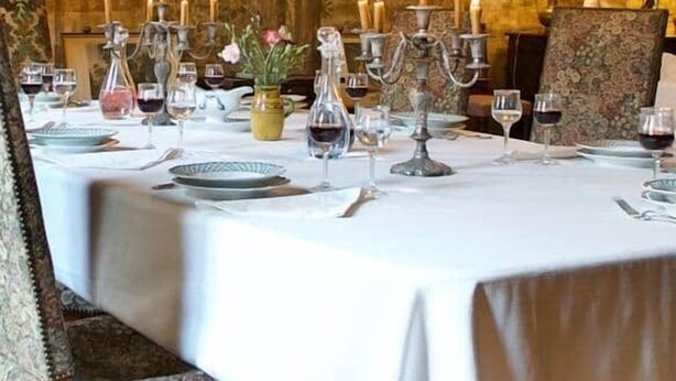 Mesa de comedor dispuesta con velas, copas y platos, rodeada de sillas y un mural con árboles.