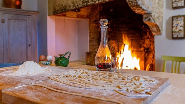 A kitchen with a wood-burning stove, a table with flour and pasta, and a green teapot.