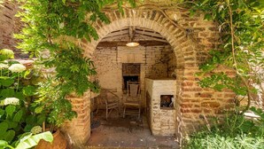 An arched brick structure with a wooden chair and a small table inside, surrounded by greenery.