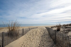 Een zandpad aan het strand met een houten hek en schaarse begroeiing.
