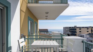 Un balcón con mesa y sillas blancas, vista al mar y un edificio con paredes exteriores azul verdoso y blanco.