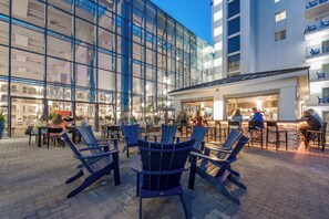 Een buitenruimte met Adirondack stoelen en tafels, een glazen gebouw met verlichting van binnen en een bar waar mensen zijn.