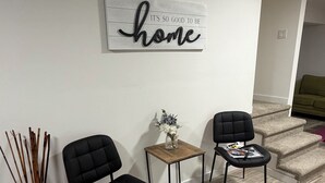 Uma sala de estar moderna com duas poltronas pretas estofadas, uma pequena mesa lateral de madeira com um vaso, e uma placa na parede com a inscrição 