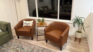 Uma sala de estar com duas poltronas de couro marrom, uma delas com uma almofada com a palavra 'hope', uma mesinha de madeira, um sofá verde, um tapete estampado e um vaso de plantas.