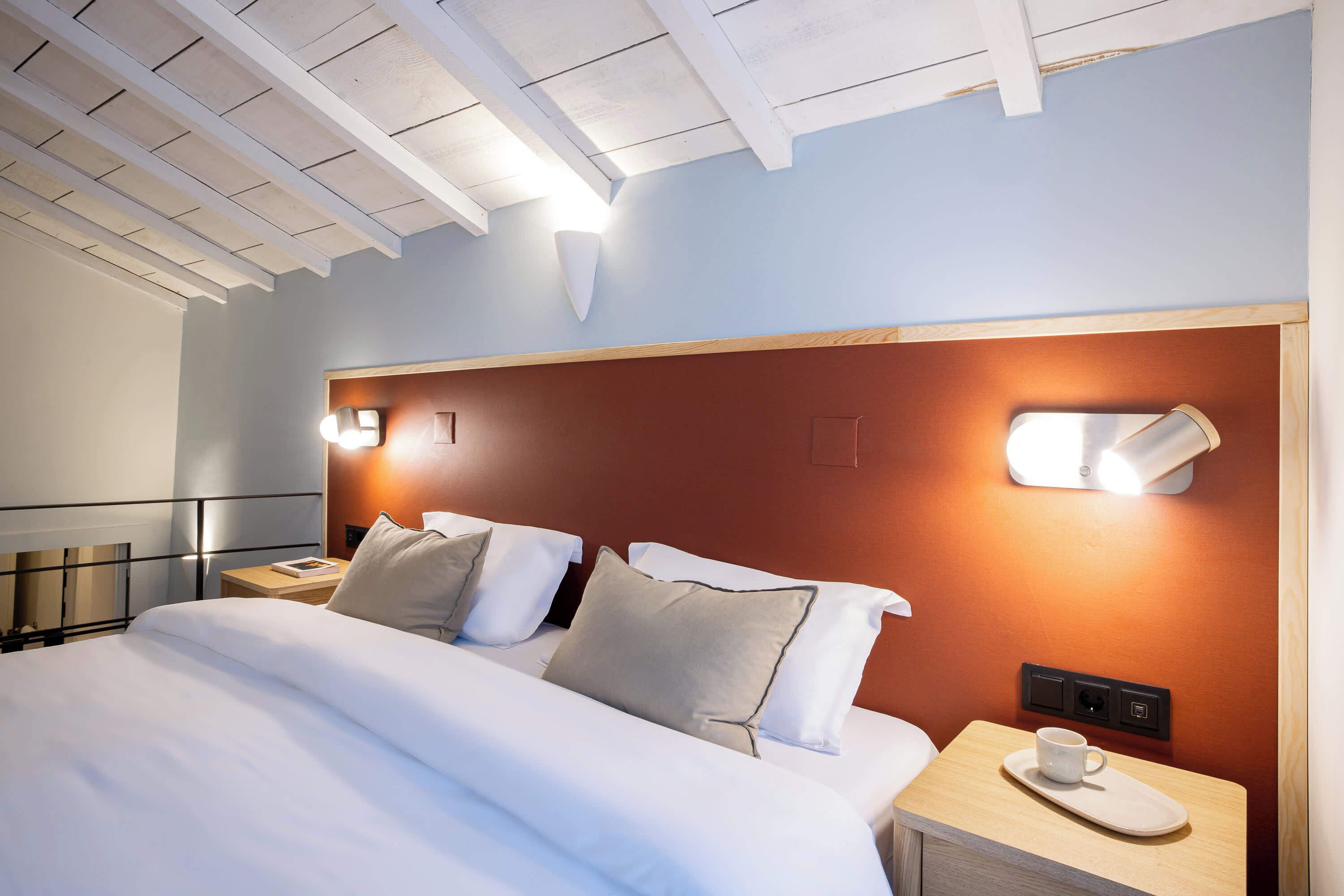 A neatly made bed with white linens and two pillows, a bedside table with a cup and saucer, and a wall-mounted light fixture.