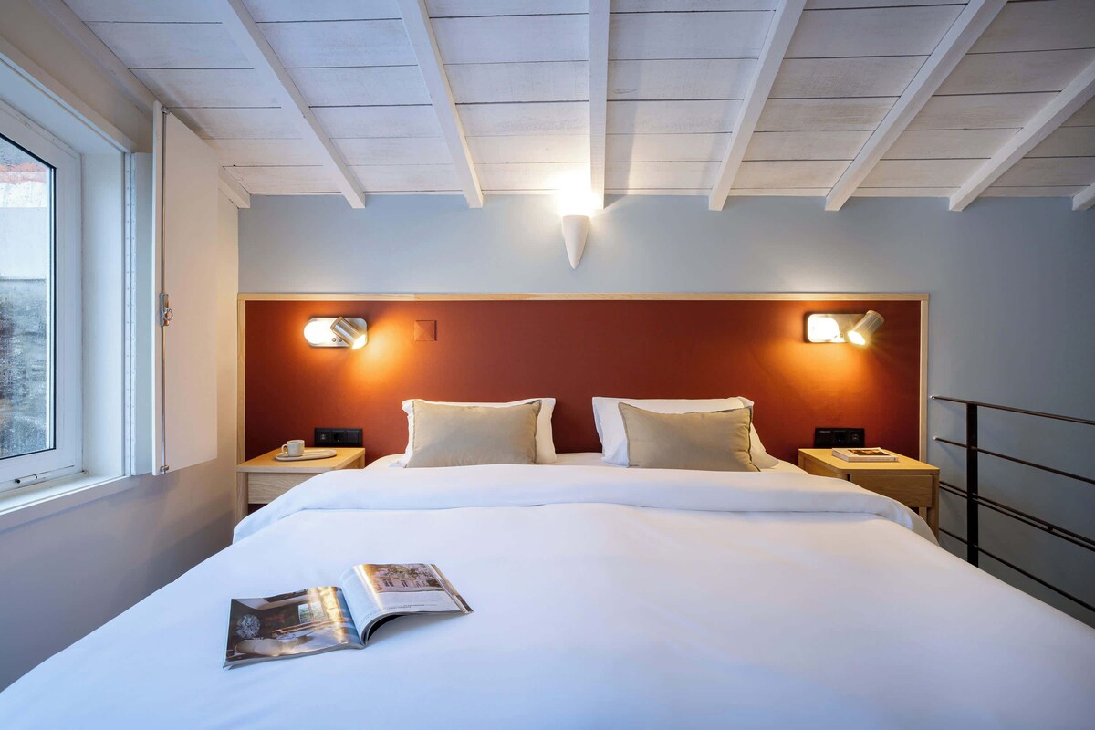 Een net opgemaakt bed met een tijdschrift erop, twee nachtkastjes met lampen, een raam en een houten plafond.