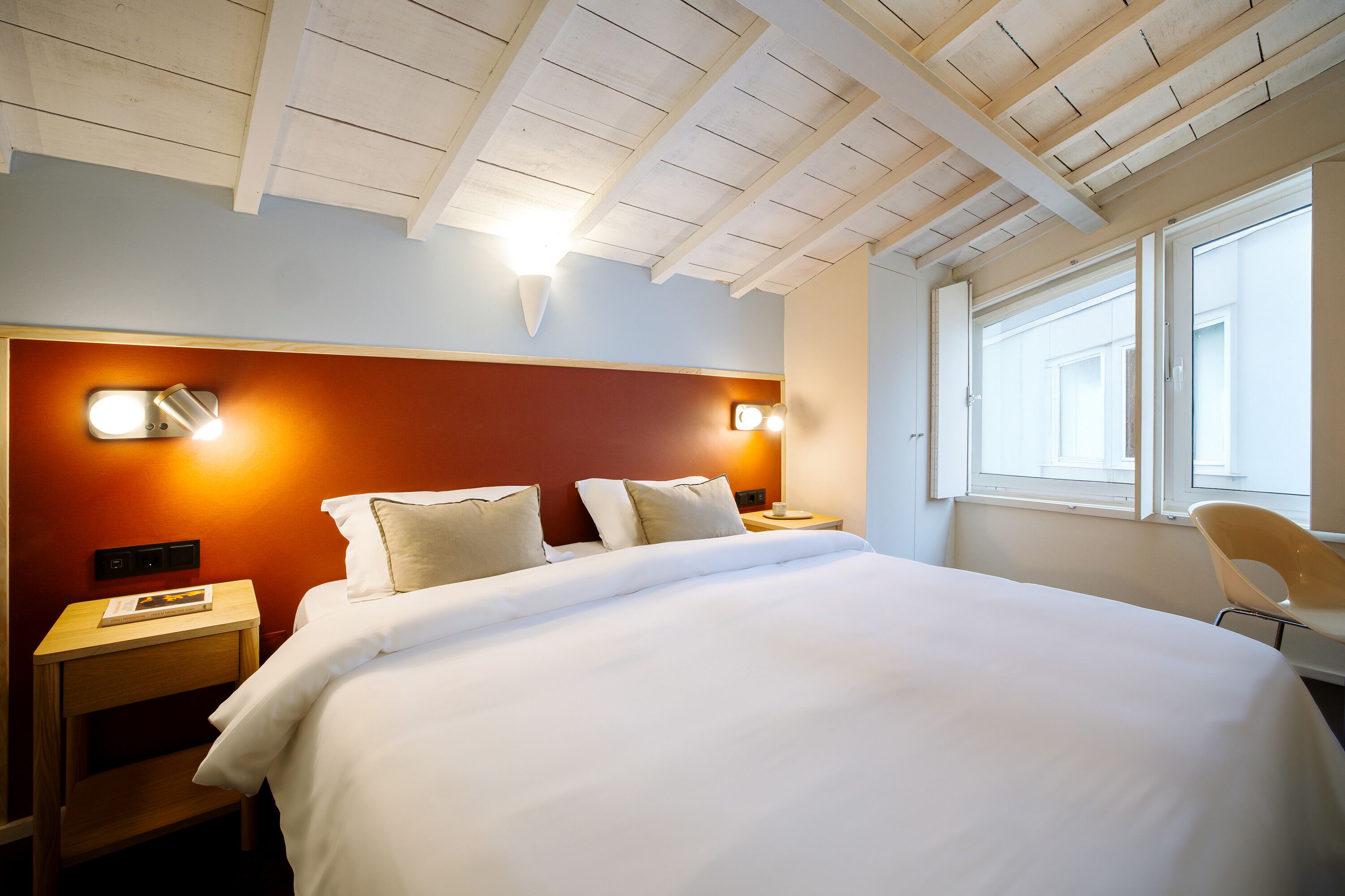 A hotel room with a wooden ceiling, a bed with white bedding, two bedside lamps, a nightstand, and a window with a view.
