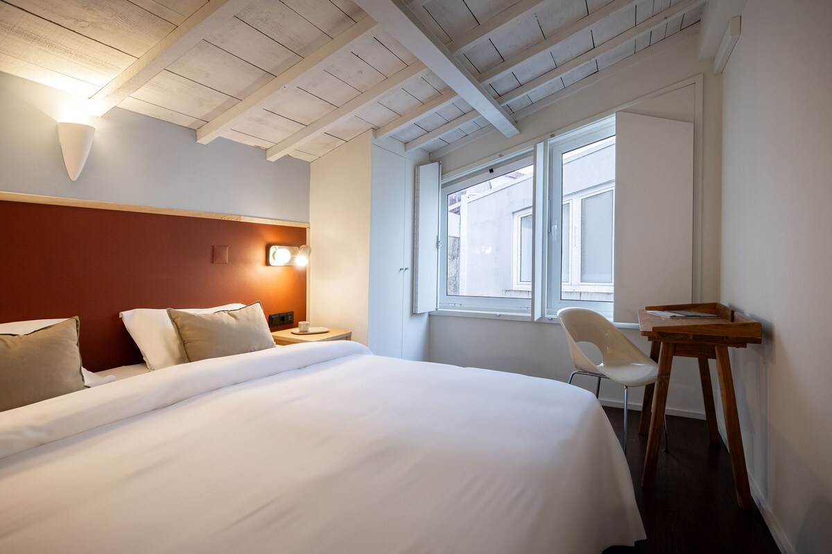 Een hotelkamer met een bed, nachtkastje, stoel, bureau en een raam met uitzicht.