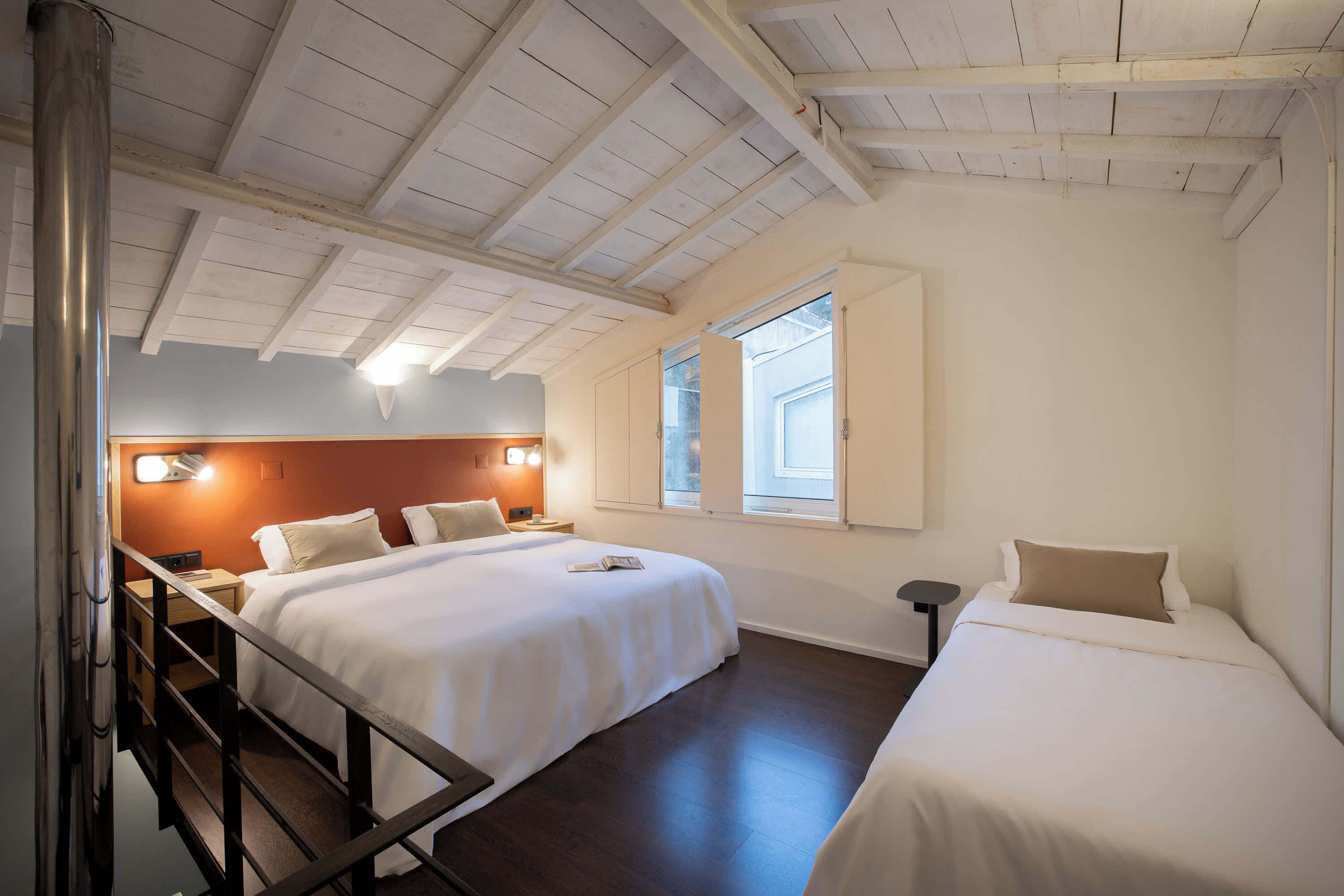A hotel room with two beds, wooden ceiling beams, a window, and a nightstand with a lamp.