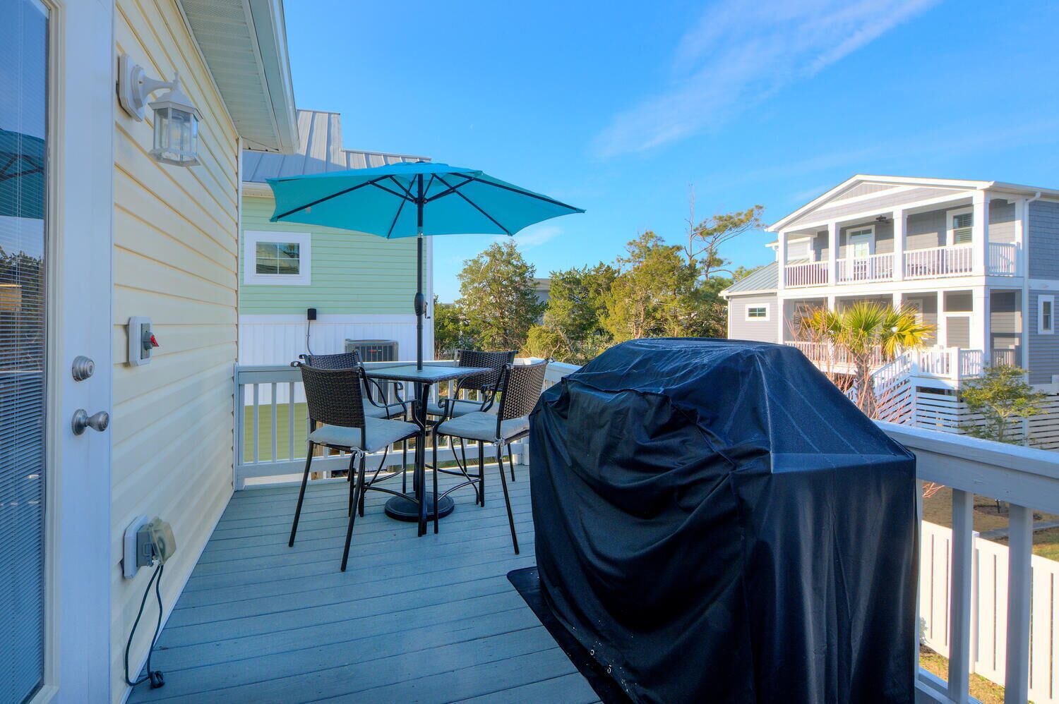 A covered outdoor deck with a table and chairs, a covered grill, and a view of neighboring buildings.