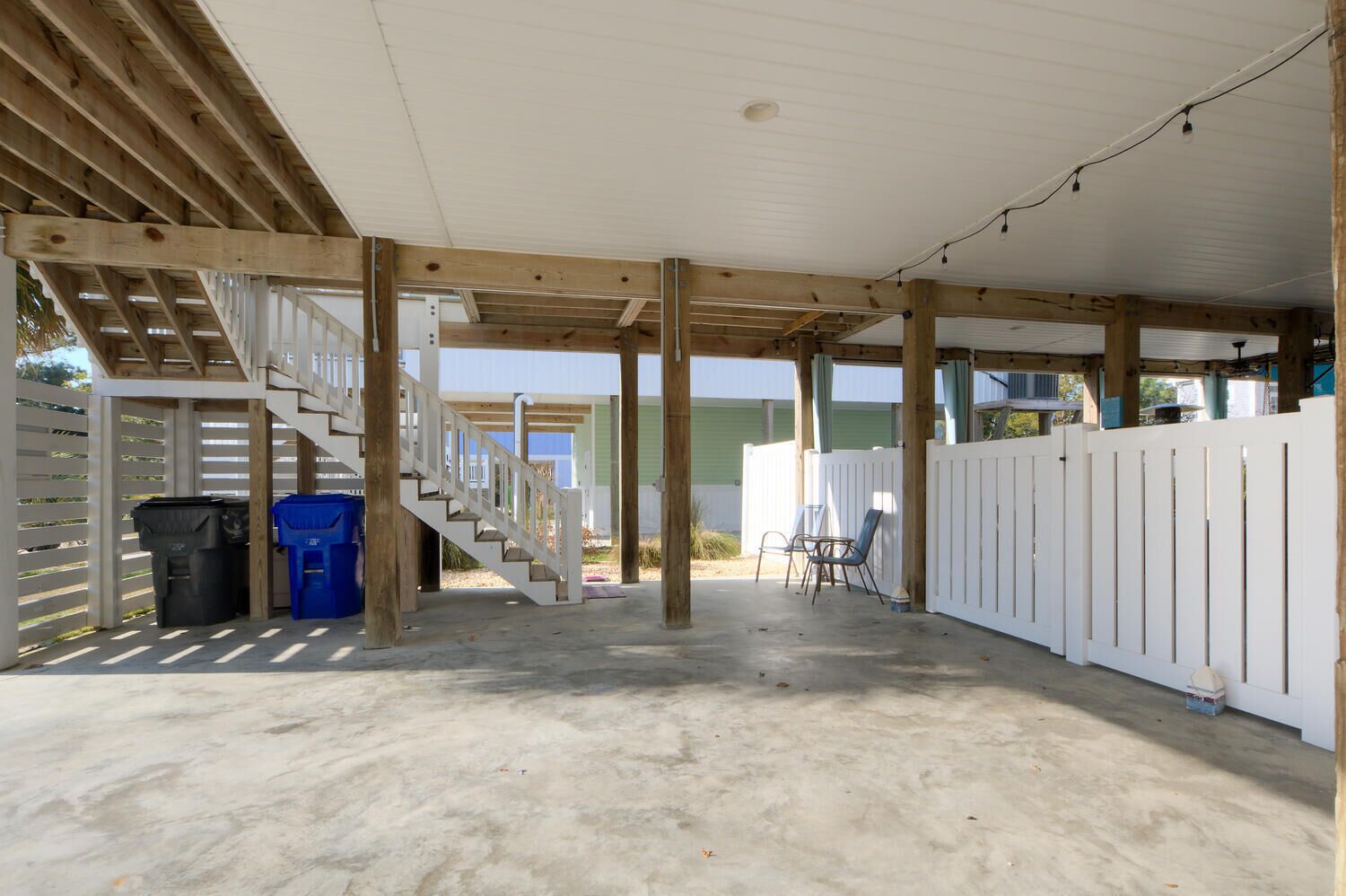 A covered outdoor area with a concrete floor, wooden beams, a staircase, and a white fence.