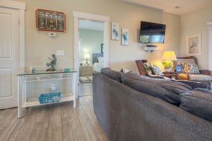 A living room with a grey sofa, a glass coffee table, a flat-screen TV, and a wall-mounted shelf with decorative items. - Experience beachside bliss at Fountains Folly, a spacious 4-bedroom home in Oak Island. (Oak Island)