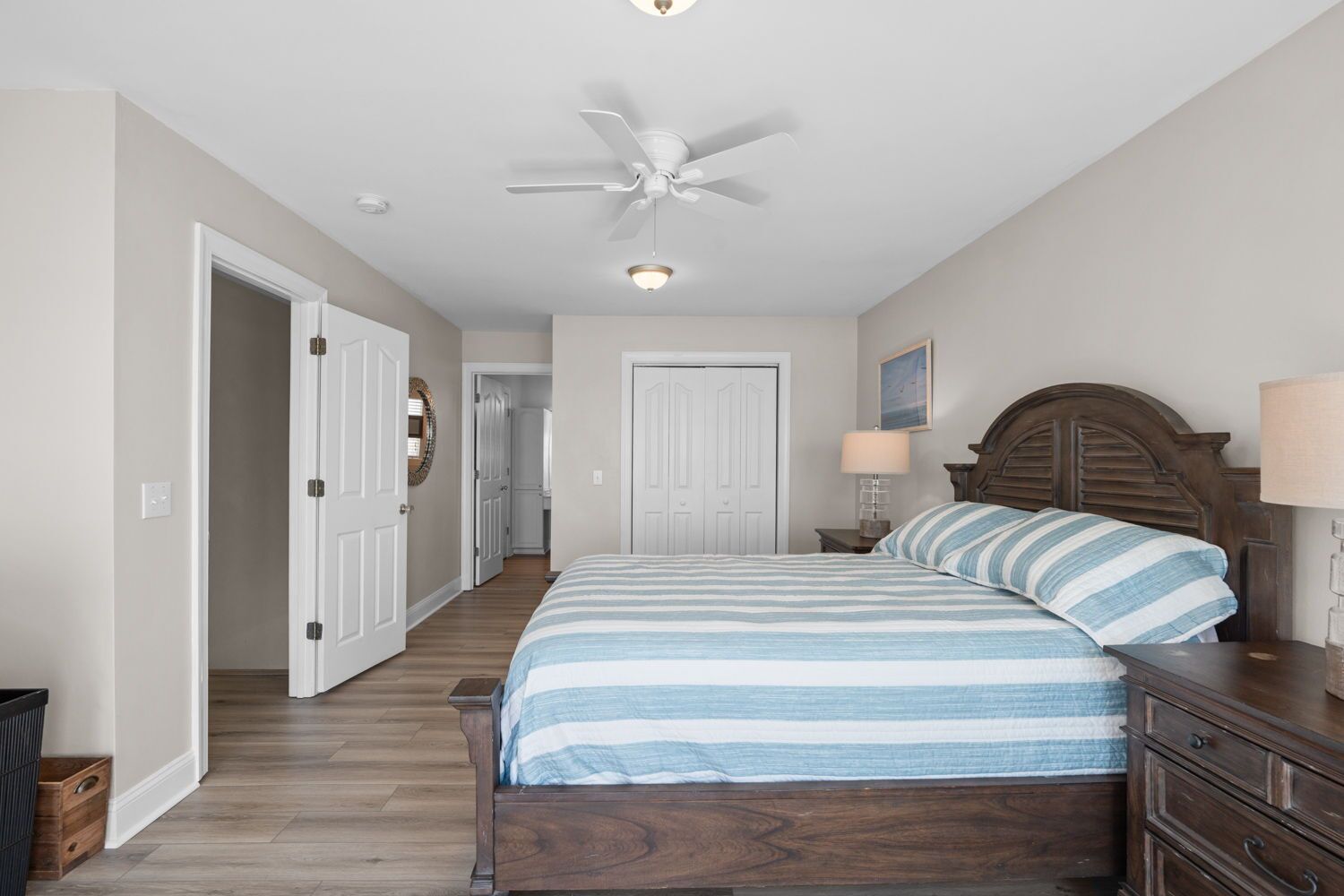 A bedroom with a bed, a nightstand, a lamp, a ceiling fan, and a door leading to another room.