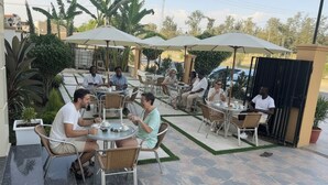 An outdoor café with tables and chairs under umbrellas, surrounded by greenery and a paved walkway. - Golden Gate Hotel (Awassa)