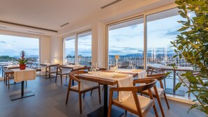 A modern dining area with large windows, wooden furniture, and a view of the cityscape. - Hotel with swimming pool in Olbia (Olbia)