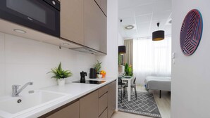 A modern kitchen with a white countertop, stainless steel sink, and a microwave. There is a dining area with a table and chairs, and a bed with a gray headboard. - Modern Studio for Two | Parking and Air Conditioning (Warszawa)